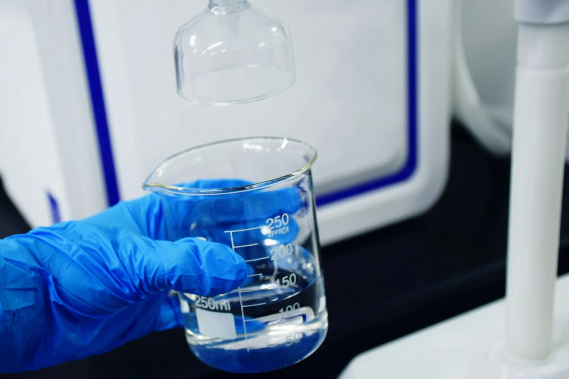 Gloved hand holding a beaker of blue solution under lab equipment